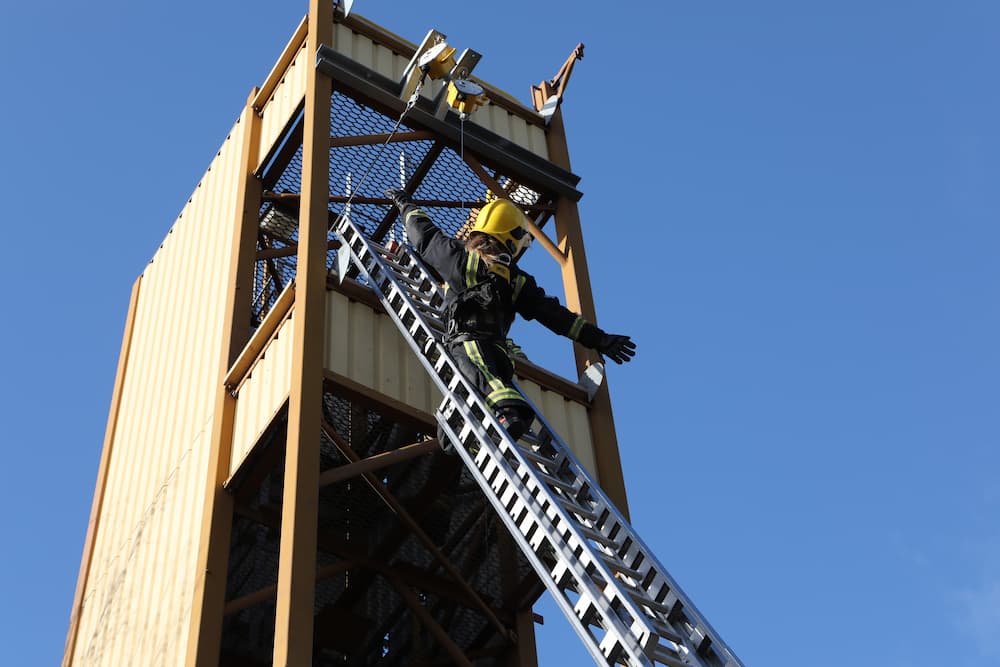 A firefighter candidate at the top of a ladder with their legs locked and releasing their arms.
