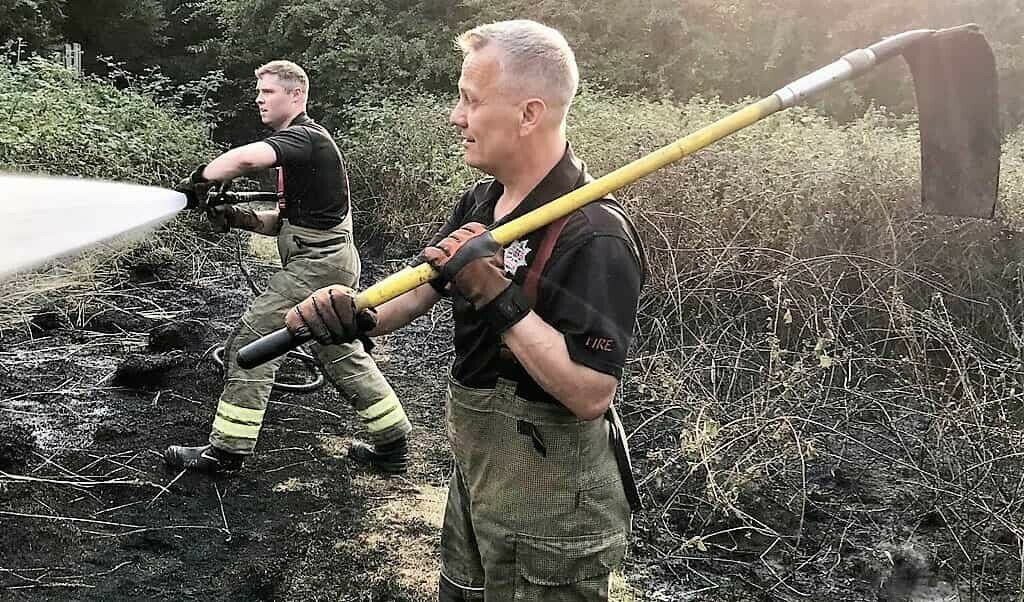 Two firefighters tackle a scrubland fire, one with a hose the other with a fire 'beater'