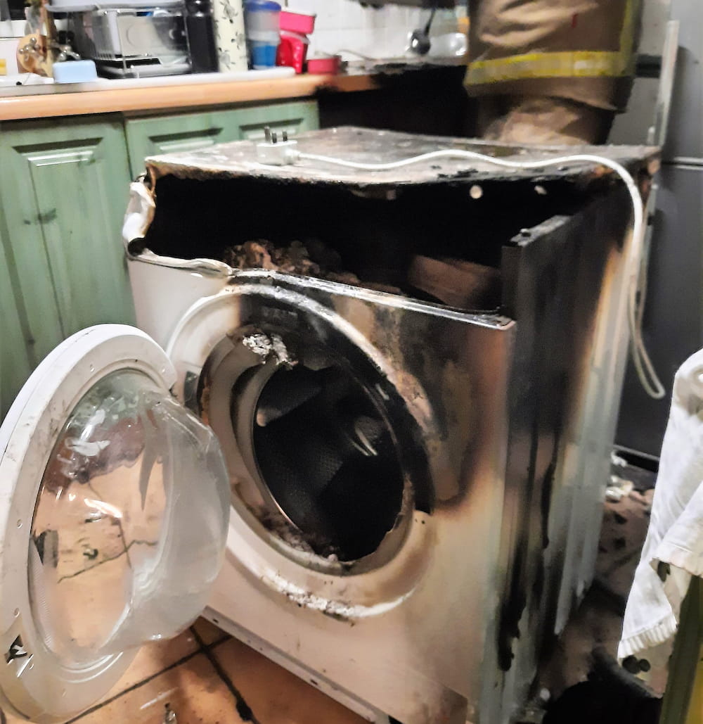 A washing machine in the middle of a kitchen, with fire damage to its top and door area