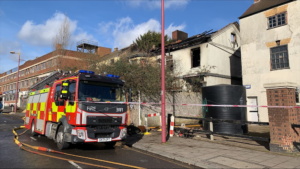 A side view of the premises with fire damage to roof, a fire engine parked next to it.