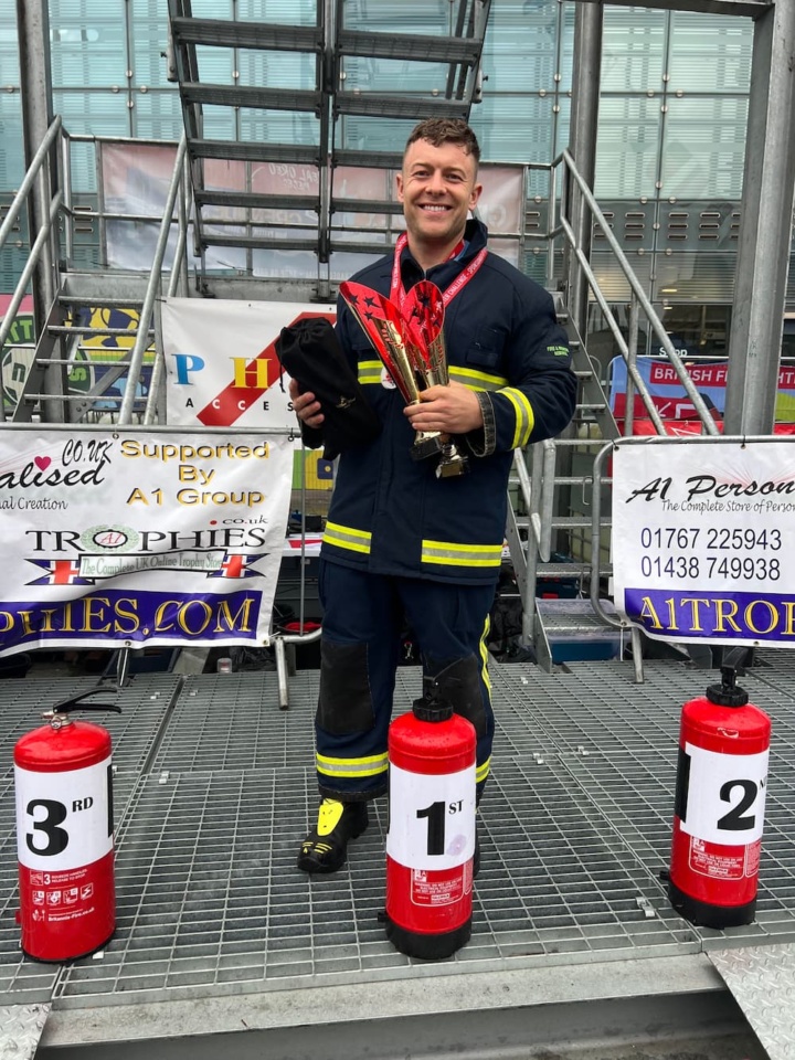 Firefighter Jack Baker holding his trophies in front of 3 fire extinguishers with 1st, 2nd and 3rd place on each one respectively.