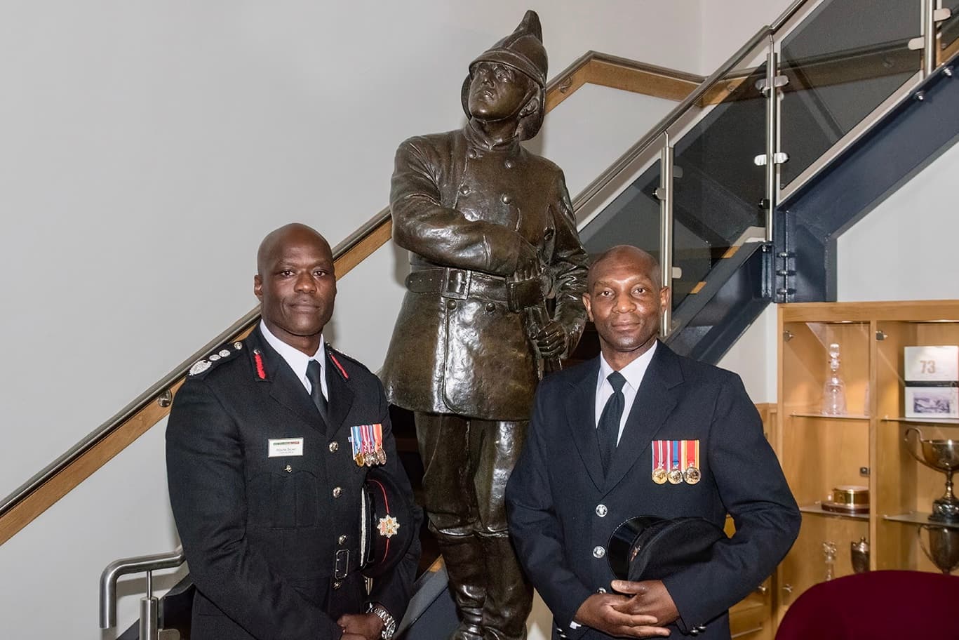 Chief Fire Officer and a firefighter, both in smart uniforms and wearing medals, stand in front of a bronze statue of a firefighter