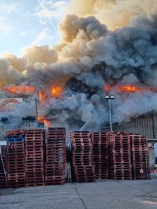 Stacks of pallets in front of large plumes of smoke and flames
