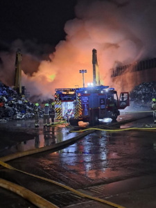 A fire engine and firefighters tackling fire, with smoke rising from a scrap pile