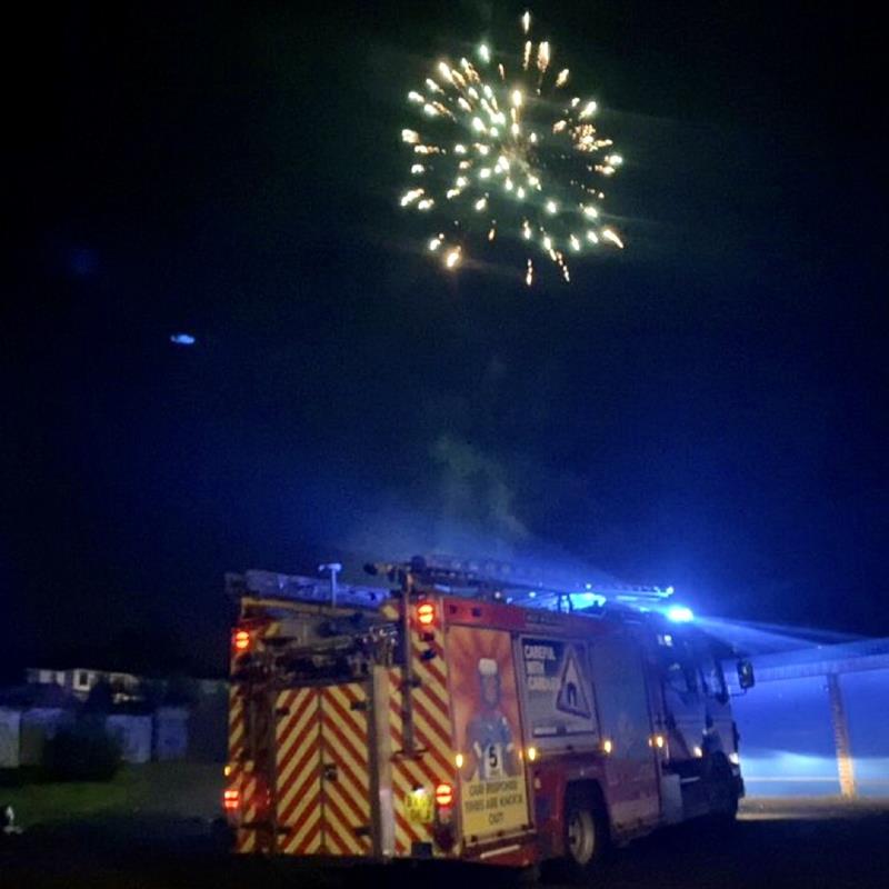 A fire engine's blue light illuminate the dark as a firework goes off in the sky above